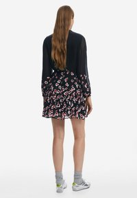 Schwarzes Blumenkleid mit langen durchsichtigen Ärmeln, gerafftem Saum, verziert mit roten und weißen Blumen, kombiniert mit grauen Socken und silbernen Schuhen.