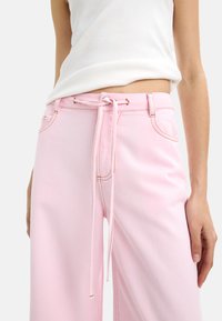 Różowe spodnie w szerokim kroju z wiązaniem w talii, z kontrastowymi szwami i dwoma przednimi kieszeniami. Materiał ma gładką, przypominającą dżins teksturę.