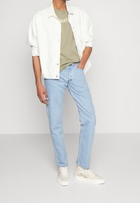 Vit denimjacka, olivgrön t-shirt med text, ljusblå jeans och krämfärgade sneakers med blommotiv.