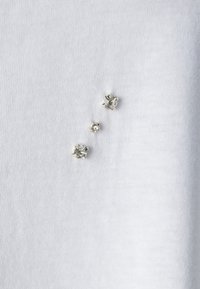 Tela blanca con tres grupos de cristales plateados decorativos de diferentes tamaños, cada uno con un diseño facetado que crea un sutil interés visual.