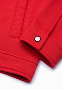 Tessuto di felpa rossa con una superficie liscia, dotato di un polsino con un bottone nero e un centro bianco a contrasto, che mostra dettagli di cucitura.