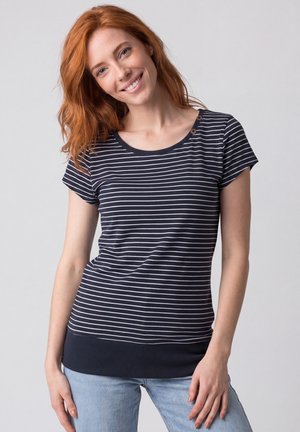 Rothaarige Frau lächelt, trägt ein marineblau-weiß gestreiftes kurzärmeliges Oberteil mit Knöpfen an der Schulter und hellblaue Jeans.