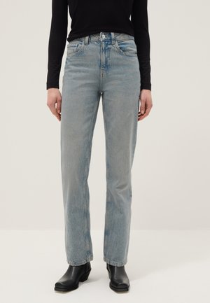 Persoon draagt lichtblauwe rechte jeans, zwarte enkellaarsjes met hak en een aansluitend zwart shirt met lange mouwen, staand tegen een effen achtergrond.