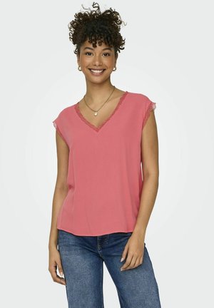 Blusa de manga corta en coral con escote en V y borde de encaje en el cuello y las mangas; tela ligera; corte holgado; combinada con jeans oscuros.