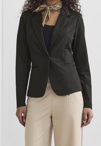 Černý blazer s klopovými límci a dvěma předními kapsami, v kombinaci s tmavým topem a béžovými kalhotami. Materiál se zdá být hladký a dobře padnoucí.