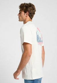 Camiseta de color crema con mangas cortas, que presenta un gráfico de montaña y atardecer en la parte posterior en tonos azules, rosas y blancos.