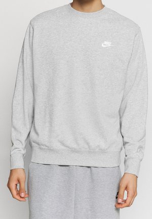 Person iført lysegrå Nike sweatshirt og matchende joggingbukser, begge med hvide Nike-logoer på brystet og låret.