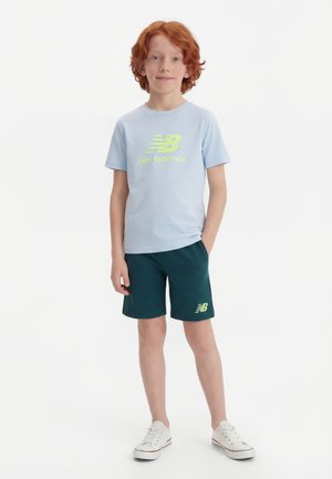 Ragazzo dai capelli rossi in piedi con una mano in tasca, indossa una maglietta New Balance azzurro chiaro, pantaloncini verde scuro e scarpe da ginnastica bianche su sfondo bianco.