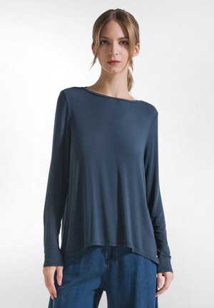 Kvinde med lyst brunt hår samlet i en lav hestehale iført en mørkeblå bluse med lange ærmer og blå jeans mod en ensfarvet hvid baggrund.