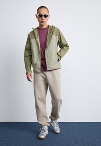 Veste légère olive verte avec capuche, assortie à un t-shirt bordeaux et un pantalon de survêtement beige. Des baskets argentées complètent la tenue.
