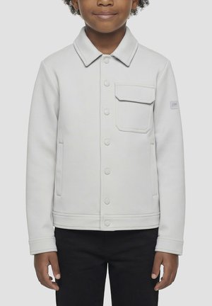 Niño con chaqueta gris claro abotonada con un bolsillo en el pecho con solapa y pantalones negros, de pie frente a un fondo liso.