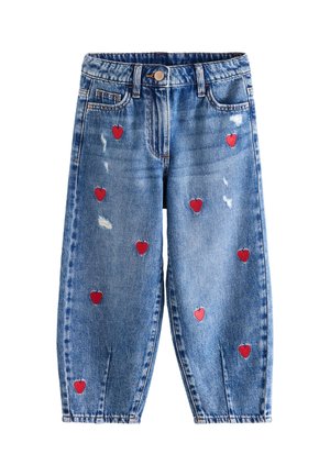Enfant portant un jean bleu ample avec des patchs en forme de cœur rouge, des chaussettes blanches, des baskets rouges et une chemise blanche, debout les mains dans les poches.