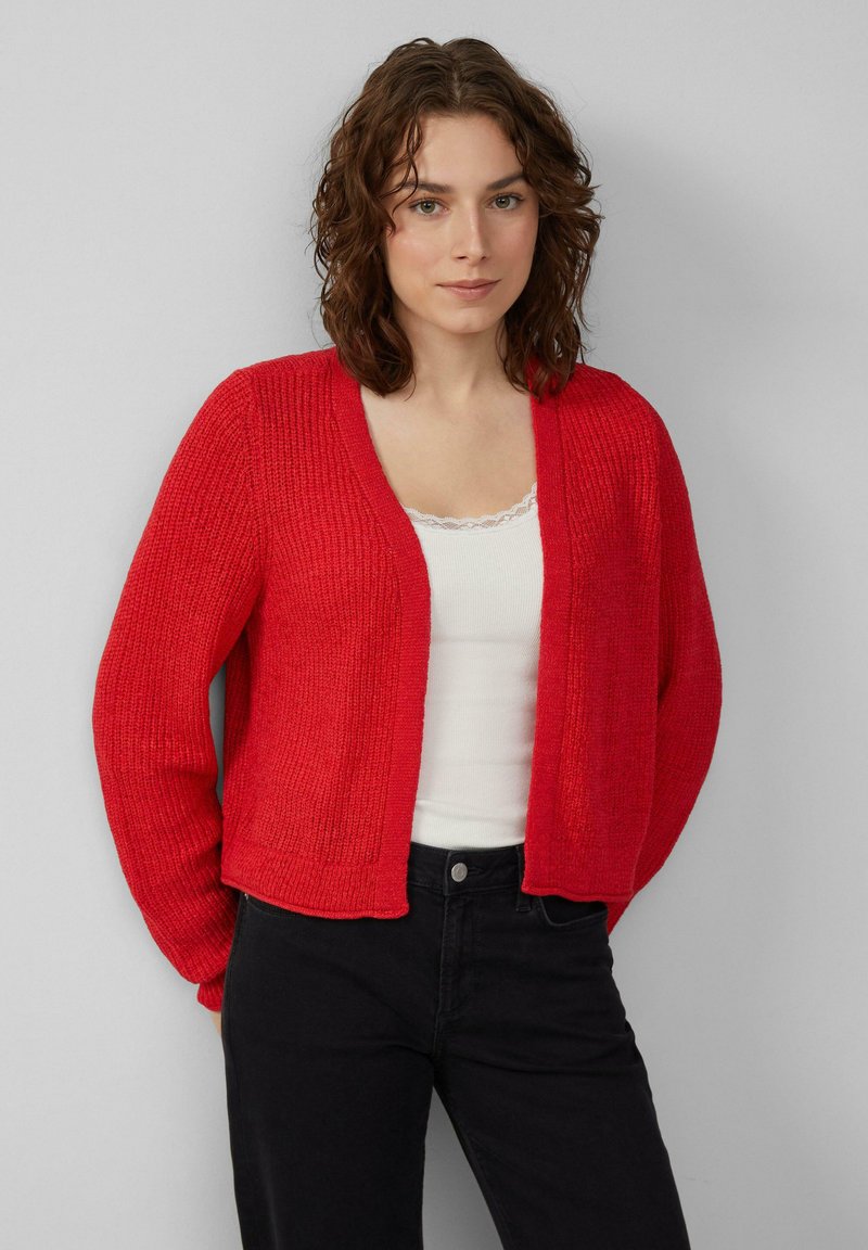 Rode gebreide cardigan met een textuurstof, open voorkant en ribbel-details. Gedragen over een witte top, gestyled met zwarte jeans.