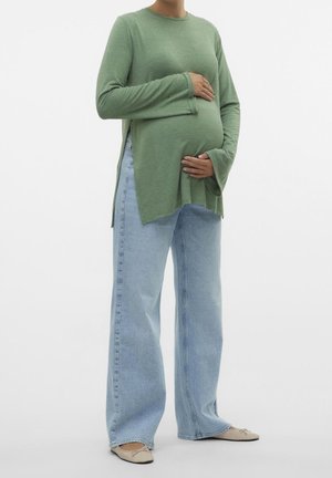 Personne enceinte portant un haut vert à manches longues et un jean bleu clair, tenant son ventre avec les deux mains, debout sur fond blanc.