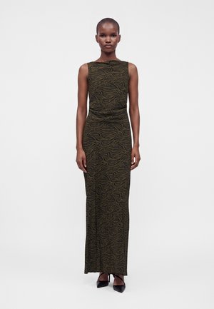 Femme portant une robe sans manches longue jusqu'au sol avec un motif ondulé vert foncé et noir, debout les bras détendus le long du corps.