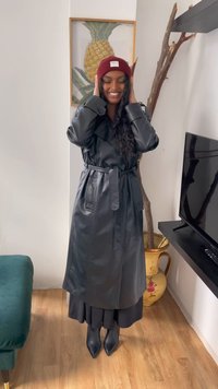 Black leather trench coat with a belted waist, worn over a long black dress and pointed ankle boots; also features a maroon beanie.