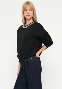 Mujer rubia vestida con un sudadera negra con un escote adornado con joyas y unos vaqueros de tiro alto azul oscuro, de pie con una mano en el bolsillo.