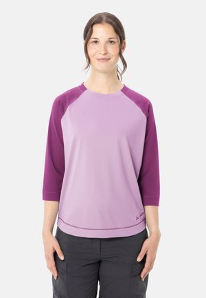 Jeune femme aux cheveux foncés portant une chemise à manches longues bicolore violet et un pantalon noir, debout devant un fond blanc uni.