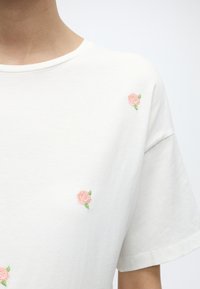 Hvit kortermet skjorte med små rosa broderte roser ved kragen, brystet og nederst foran.