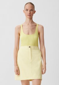 Frau in einem blassgelben, gerippten Tanktop und einem passenden, hoch taillierten Rock mit Knopfdetail, vor einem schlichten hellen Hintergrund stehend.