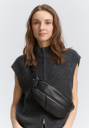 Borsa a tracolla nera in pelle con forma curva, dotata di chiusura con zip e tracolla regolabile. Indossata sopra un top in maglia grigio scuro.