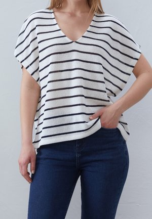 Vrouw draagt een losse witte gebreide top met zwarte horizontale strepen en donkerblauwe jeans, één hand in de zak, tegen een effen achtergrond.
