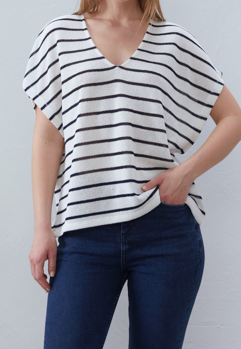 Femme portant un haut en maille blanc ample à rayures horizontales noires et un jean bleu foncé, une main dans la poche, sur un fond uni.