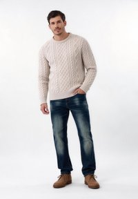 Homme debout en pull beige en maille torsadée, jean bleu et chaussures en suède marron, une main dans la poche, sur fond blanc uni.