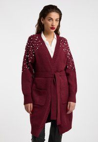 Cardigan tricotat în culoarea burgund, cu talie accentuată printr-un brâu, având accente din perle pe umeri, buzunare adânci și mansete cu model ribat.