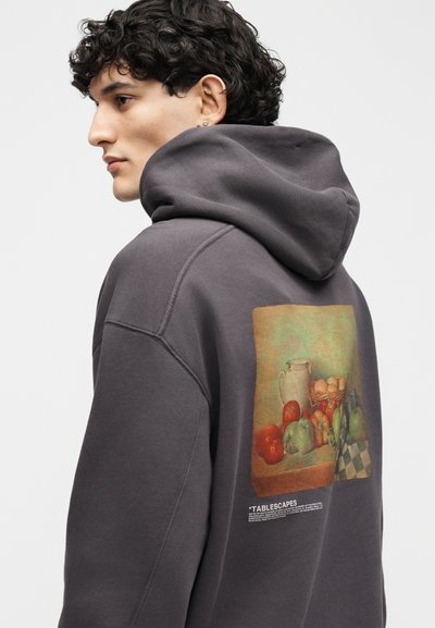 Jaunas vyras su garbanotais plaukais, dėvintis tamsią gobtuvinę su spauda gale, kurioje pavaizduotas vaisių, kiaušinių ir ąsočio nuotraukos atvaizdas.