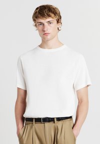 T-shirt bianco a maniche corte in maglia con scollatura rotonda, caratterizzato da un tessuto testurizzato. Abbinato a pantaloni beige plissettati e una cintura nera.