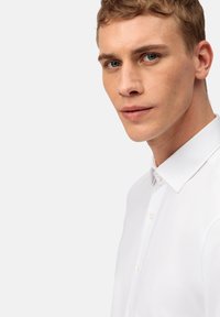 Chemise blanche avec un col structuré, fermeture à boutons sur le devant et une texture lisse ; modèle montré de côté avec un fond neutre.