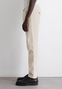 Beige cotton trousers with a straight cut, side pockets, back pockets, and a single button at the waist. Paired with black loafers.