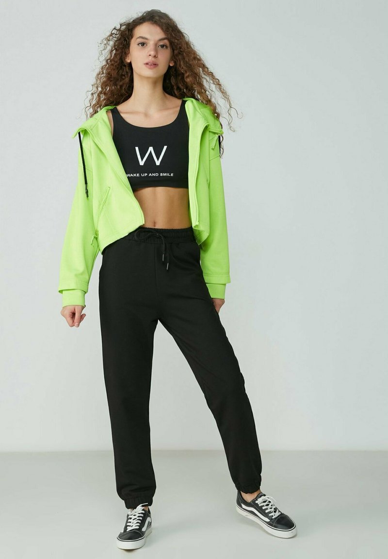 Neon-grüner verkürzter Hoodie, schwarzes Slogan-Crop-Top, schwarze tapered Sweatpants und schwarze Sneakers mit weißen Akzenten. Glatter Stoff, lässiges Design.