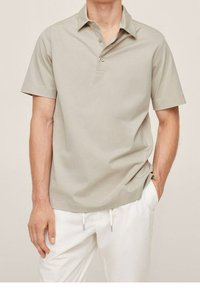 Světle béžová polo košile s krátkým rukávem a límcem, obsahující tři knoflíky a volný střih, spárovaná s bílými kalhotami.