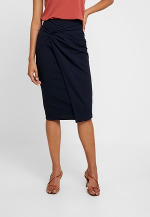 Femme portant une jupe portefeuille bleu marine longueur genou, un top sans manches couleur rouille et des sandales à talons hauts rouges à brides, debout avec la main sur la hanche.