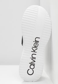 Černé tenisky s bílou gumovou podrážkou, s texturovaným gripem a logem "Calvin Klein" v tučném černém písmu.