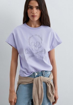Mujer que lleva una camiseta lila clara con un diseño de calavera y huesos cruzados, vaqueros azules y un suéter beige atado alrededor de su cintura.