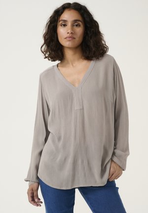 Long-sleeve, light gray top with a V-neck and subtle textured fabric. Curved hem, relaxed fit, paired with blue jeans.