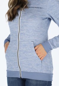 Light blue zip-up hoodie with a textured finish, featuring side pockets, ribbed cuffs, and a logo on the upper left.