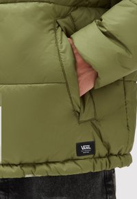 Chaqueta acolchada de color verde oliva con un bolsillo lateral y forro rosa. Presenta puños elásticos y una etiqueta negra de Vans en el dobladillo.
