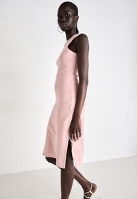 Knit, sleeveless pink dress with a fitted shape, side slit, and ribbed texture. Model stands in profile against a neutral background.