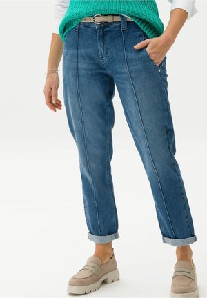 Jean en denim bleu clair avec une coupe droite, des poignets retroussés, des coutures visibles et des poches latérales, accompagnés de chaussures beige.