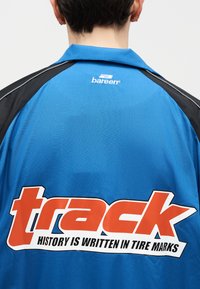 Άτομο που φοράει μπλε και μαύρο μπουφάν με το λογότυπο "bareen" στο πάνω μέρος της πλάτης και μεγάλο κείμενο "track" με το σύνθημα "Η ιστορία γράφεται στις σημάδια ελαστικών."