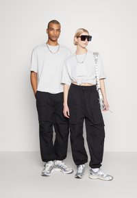 Szürke, oversize pólók, fekete cargo nadrág zsebekkel, ezüst színű sneakers, valamint kiegészítők, mint például nagy napszemüveg és láncnyaklánc.