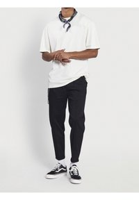 T-shirt crema a maniche corte, pantaloni neri affusolati, sneakers bianco e nere, e una sciarpa nera a fantasia attorno al collo.