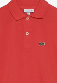 Red polo shirt made of textured fabric, featuring a classic collar, two buttons, and a green embroidered crocodile logo on the left chest.