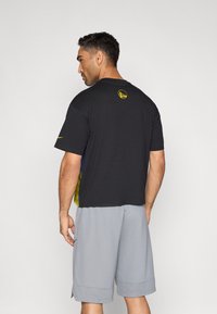 Μαύρο κοντομάνικο μπλουζάκι με κίτρινο λογότυπο στο πίσω μέρος, σε συνδυασμό με γκρι σορτς, με άνετη εφαρμογή και μαλακή υφή.