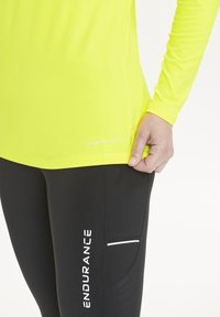 Lys gul langærmet skjorte med "ENDURANCE" trykt på kanten, parret med sorte leggings med en lomme og hvid detaljer.