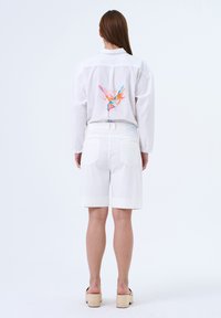 Witte lange mouwen shirt met een kleurrijk vogelontwerp op de rug, gecombineerd met witte shorts en beige platform sandalen.